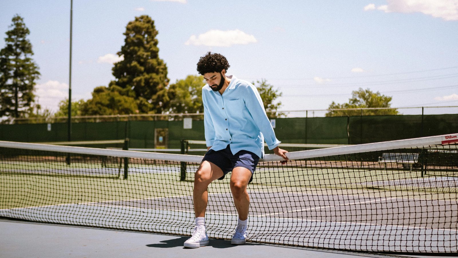 A man with curly hair sits on a tennis net at an outdoor court in Bakersfield, CA.